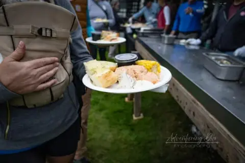 A person wearing a baby carrier holds a plate of food near a buffet table.