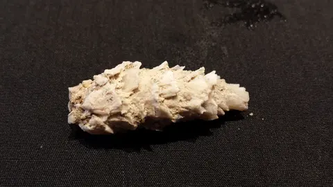 A close-up photograph of a rough white crystal or mineral specimen resting on a black textured surface.