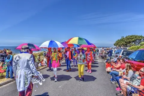 People walking in an outdoor parade carrying colorful umbrellas along a coastal road.