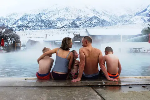A family sits on the edge of a steaming pool with snow-covered mountains in the background.
