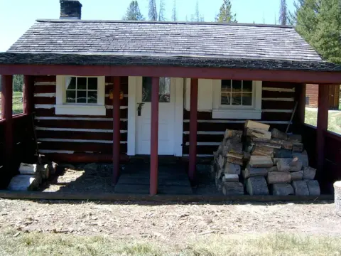An exterior view of a rustic log cabin with a covered porch and stacks of firewood.