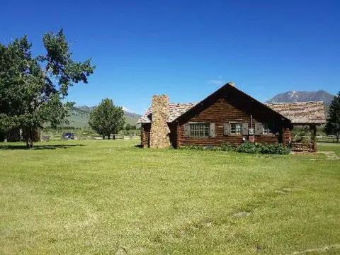 A log cabin sits in a grassy field with mountains in the background.