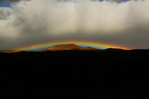 A rainbow arches across the sky above a flat-topped mountain and dark rolling hills.