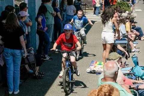 Children ride bicycles along a street past spectators watching from the sidewalk.