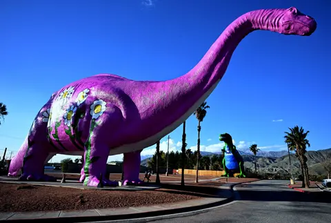A giant pink dinosaur statue painted with flowers stands outdoors against a blue sky.