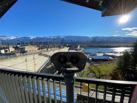 A viewing binocular overlooks a dam with snowy mountains in the background.