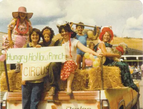 People in costumes ride on the back of a truck holding a circus sign.