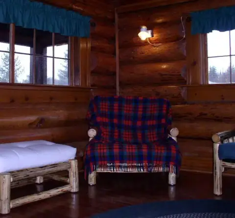An interior view of a rustic log cabin with windows, log walls, and armchairs.
