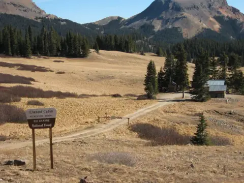 A dirt road leads through a mountain valley toward a cabin with a sign in the foreground.