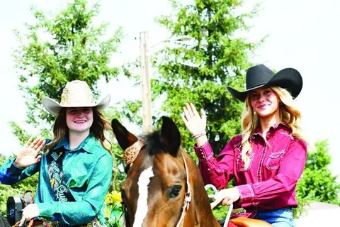 Two young women in cowboy hats wave while riding a horse outdoors.