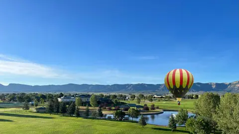 A hot air balloon floats above a green park with mountains in the distance.