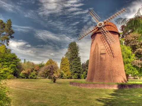 A wooden windmill stands in a grassy park surrounded by trees.