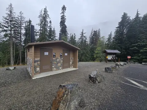 An outdoor public restroom building stands in a gravel parking area surrounded by tall evergreen trees.