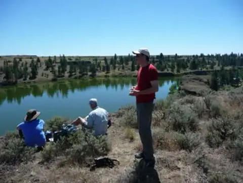Three people stand and sit on a hillside overlooking a body of water.
