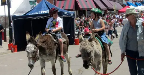 Two children wearing helmets ride donkeys down a street while people watch in the background.