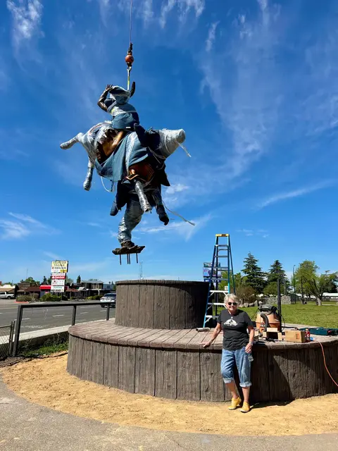 A woman stands beside a large round platform as a wrapped sculpture of a rider on a horse is lifted into the air by a crane.