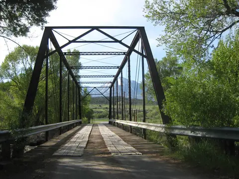 A rustic metal truss bridge spans across a rural landscape with trees and mountains in the distance.