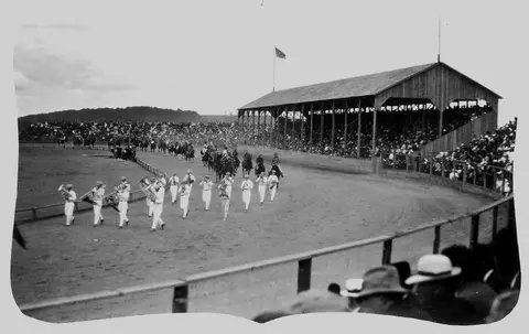 A historical photograph showing a band and horse riders in a large arena filled with spectators.