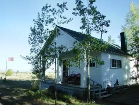 A small white building with a porch is surrounded by trees and a clear sky.