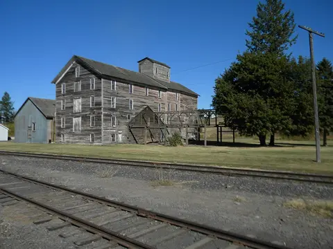 An old wooden mill stands next to railroad tracks under a clear blue sky.
