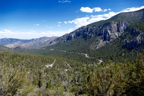 A scenic view of mountains, forests, and a winding road under a blue sky with clouds.