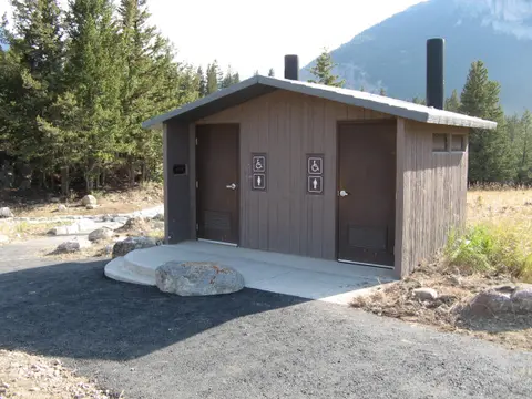 An exterior view of a brown restroom building with two doors and accessible signage surrounded by trees and mountains.