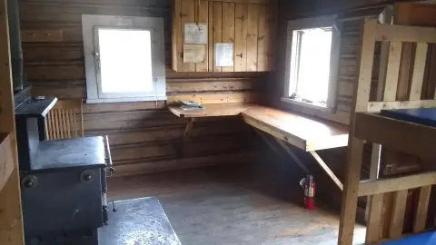 An interior view of a rustic log cabin with a wood stove, desk, cabinets, and bunk beds.