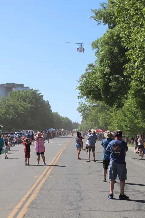 A crowd of people watch a helicopter fly down a closed street on a sunny day.