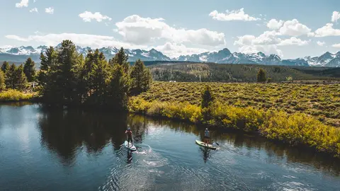 Two people paddleboarding on a scenic river with mountains in the background.