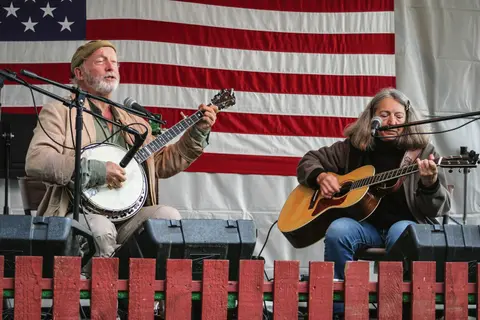 Two musicians perform with a banjo and a guitar in front of an American flag.