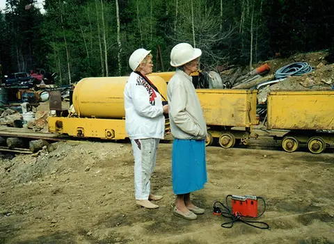 Two women wearing hard hats stand near mining equipment.