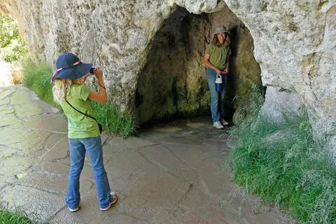 A young girl takes a photo of a woman standing in a rocky cave opening.