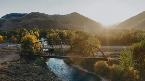 A bridge crosses a river surrounded by trees and mountains at sunset.
