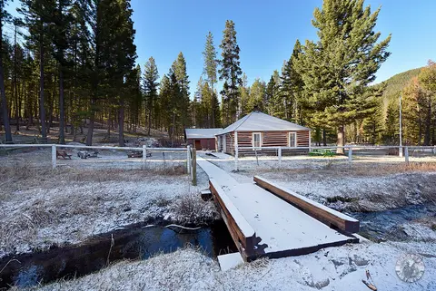 A wooden bridge leads over a stream to a log cabin surrounded by trees and snow.