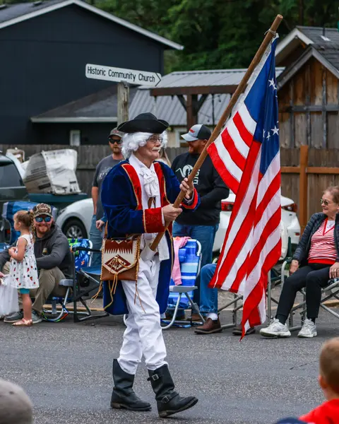 A person in a historical costume carries an American flag while walking past a crowd.