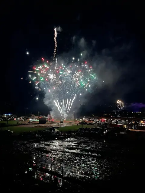 Fireworks light up the night sky over a field with cars and spectators.