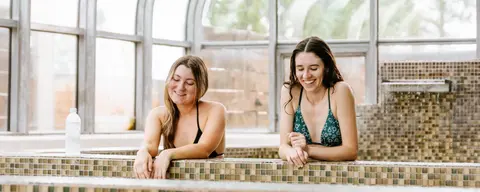Two smiling women in swimsuits relax in a tiled hot tub indoors.