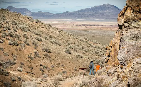 Two hikers walk on a trail through a desert landscape with hills and mountains.