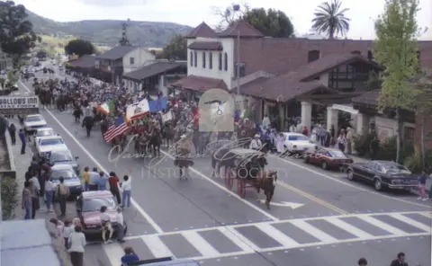 A parade with horses and flags moves down a street lined with buildings and parked cars.