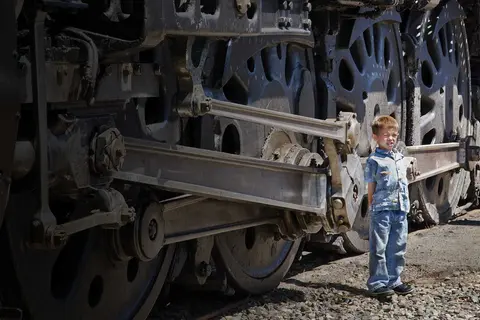A young boy stands next to the large wheels of a steam locomotive.