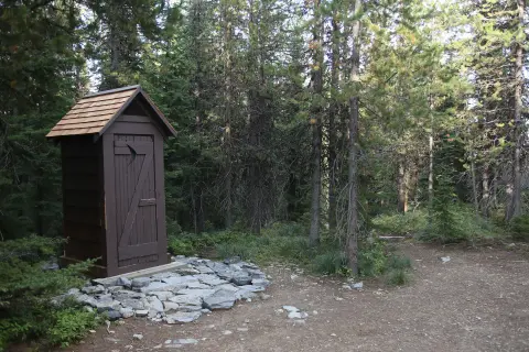 A wooden outhouse stands in a forest next to a dirt path.