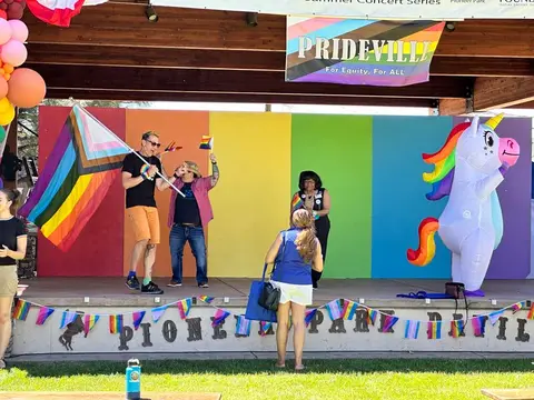 People celebrate on an outdoor stage decorated with pride flags and a rainbow backdrop.