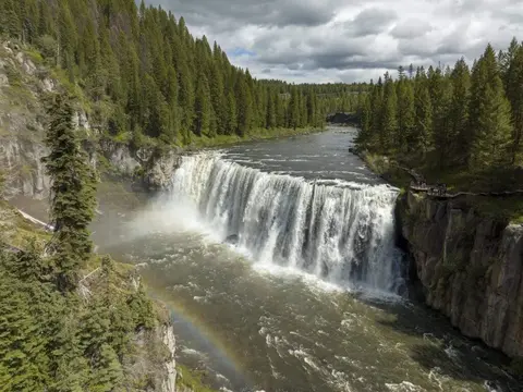 A wide waterfall flowing through a forested canyon with a rainbow visible in the mist.
