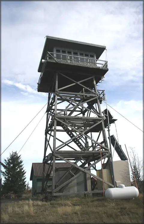 A tall wooden fire lookout tower stands outdoors under a cloudy sky.