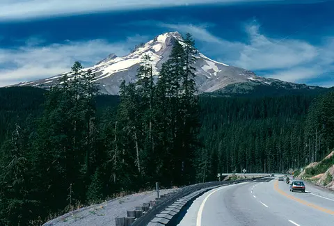 A snow-capped mountain rises above a forest and a winding highway with cars.