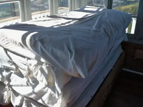 A stack of mattresses covered with white sheets inside a room with windows overlooking a landscape.