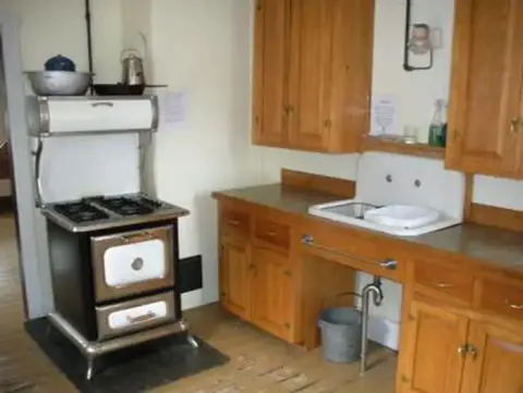 A photo showing a vintage kitchen interior with a wood stove and wooden cabinets.