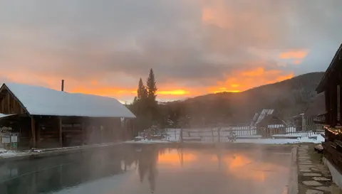A steaming pool outdoors at sunrise with snow on the ground and buildings.