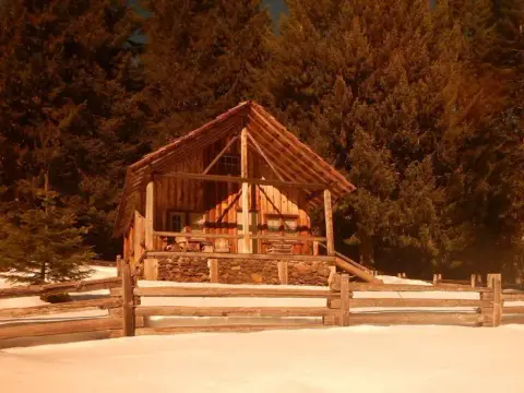 A rustic wooden cabin sits behind a wooden fence in a snowy, wooded area.