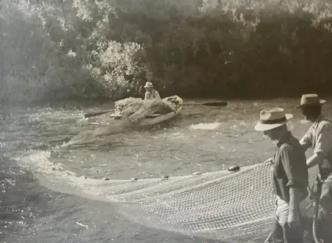 Historic photo showing people fishing with a net from a boat and the shore.